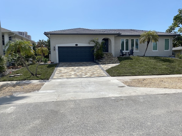 A single-story house with a dark blue garage door, a front porch with a small table and chairs, and a palm tree in the front yard.