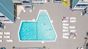 An aerial view of a rooftop pool area with a rectangular pool, lounge chairs, and a table with umbrellas.
