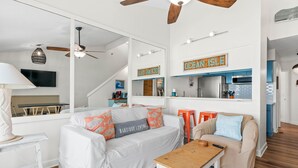 A modern living room with a white sofa, a wooden coffee table, and a beige sofa. There is a kitchen area with blue cabinets and a sign that reads "OCEAN ISLE".