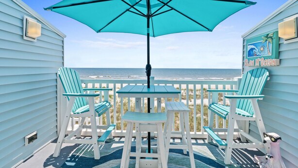 A balcony with turquoise chairs and a table, overlooking the ocean.