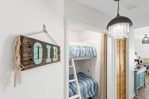 A bunk bed with a blue and white striped bedspread, a wooden sign with the letters "OIB" on the wall, and a chandelier hanging above. - Charming Oceanfront 2BR Condo + Pool on Ocean Isle Beach, NC (Ocean Isle Beach)