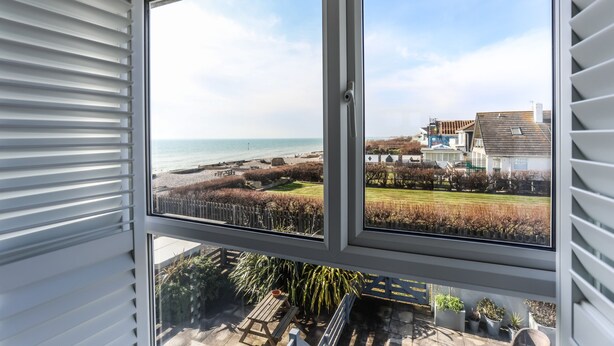 A window with a view of a beach, a grassy area, and a building in the background. - Sea View 3 Bedroom Seaside Property (Chichester)