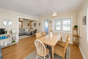 A dining area with a wooden table and chairs, a coffee station, and a living room with a sofa and a clock.