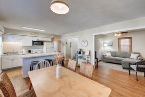 A modern kitchen with white cabinets, a dining area with a wooden table and wicker chairs, and a living room with a grey sofa and a ceiling fan.