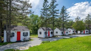 A row of small, white cabins with red doors and black shutters, surrounded by trees and a grassy area. - 14 1Br Cottage in Bar Harbor Open Hearth Inn (Trenton)
