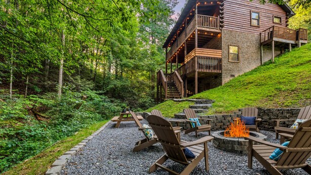 Una cabaña de troncos con base de piedra, una terraza de madera y una zona de fogata con sillas Adirondack.