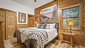 Un dormitorio de cabaña de madera con una cama, una mesita de noche, una lámpara y un cuadro de un bosque.