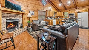 Un acogedor interior de cabaña con chimenea de piedra, asientos de cuero, una zona de cocina y un televisor grande montado en la pared.