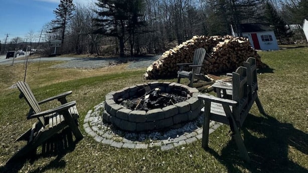 Un foyer circulaire en pierre, entouré de rondins de bois et de chaises Adirondack, installé dans un espace herbeux avec des arbres en arrière-plan.