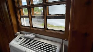 Une pièce dotée d’une grande fenêtre, d’un climatiseur blanc et offrant une vue sur une maison et des arbres.