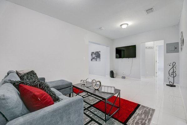 Un salon moderne avec un canapé gris, des coussins rouges et noirs, une table basse en verre et un tapis rouge et gris.