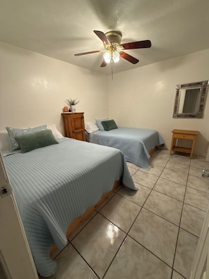 Two single beds with blue bedspreads and matching pillows, a ceiling fan, and a nightstand with a mirror.