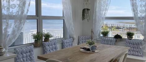 A dining area with a wooden table and white wicker chairs, large windows with sheer curtains, and potted plants on the windowsill.