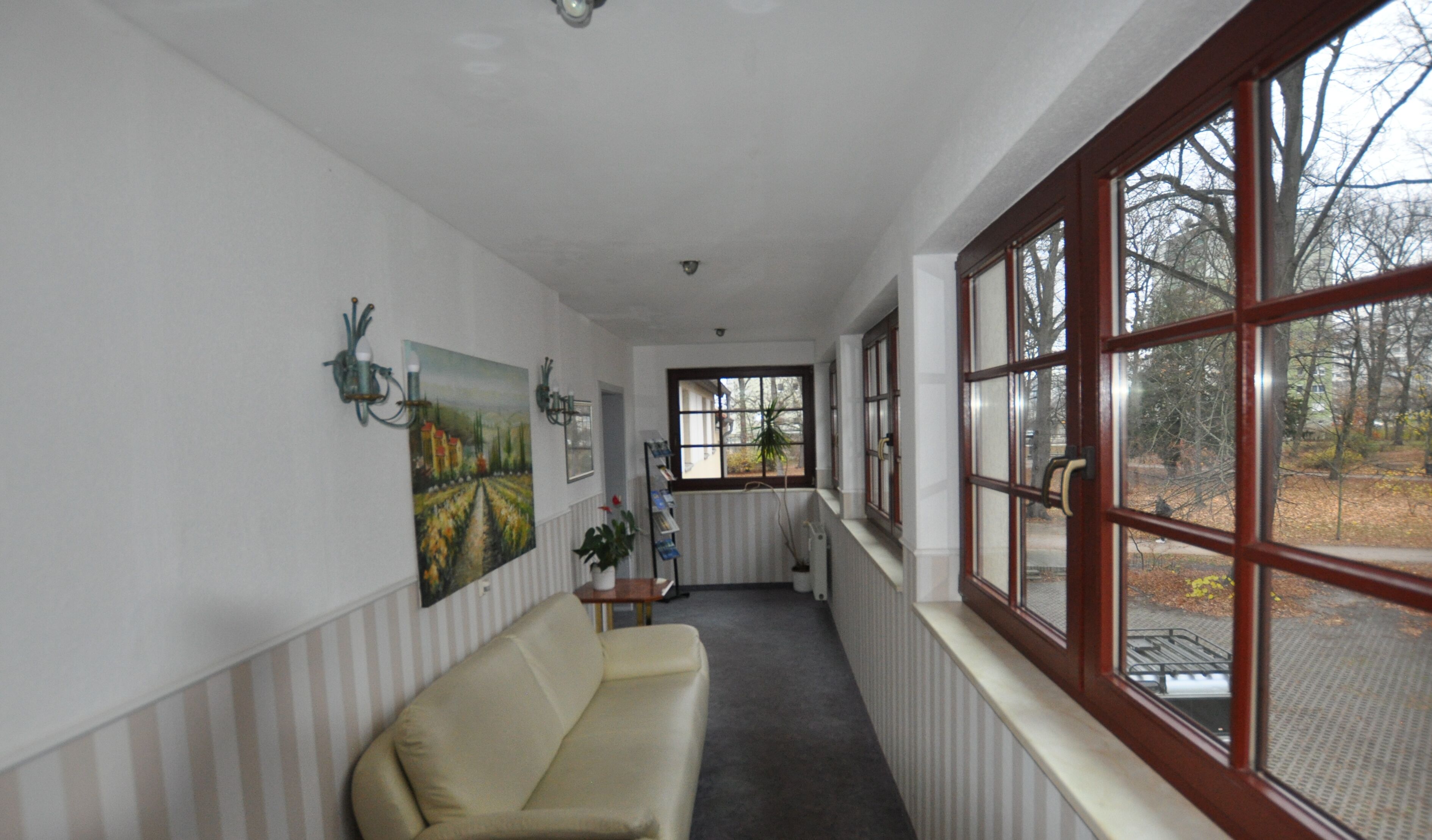 A corridor with a beige sofa, a radiator, a window with a view of trees, and a painting on the wall.