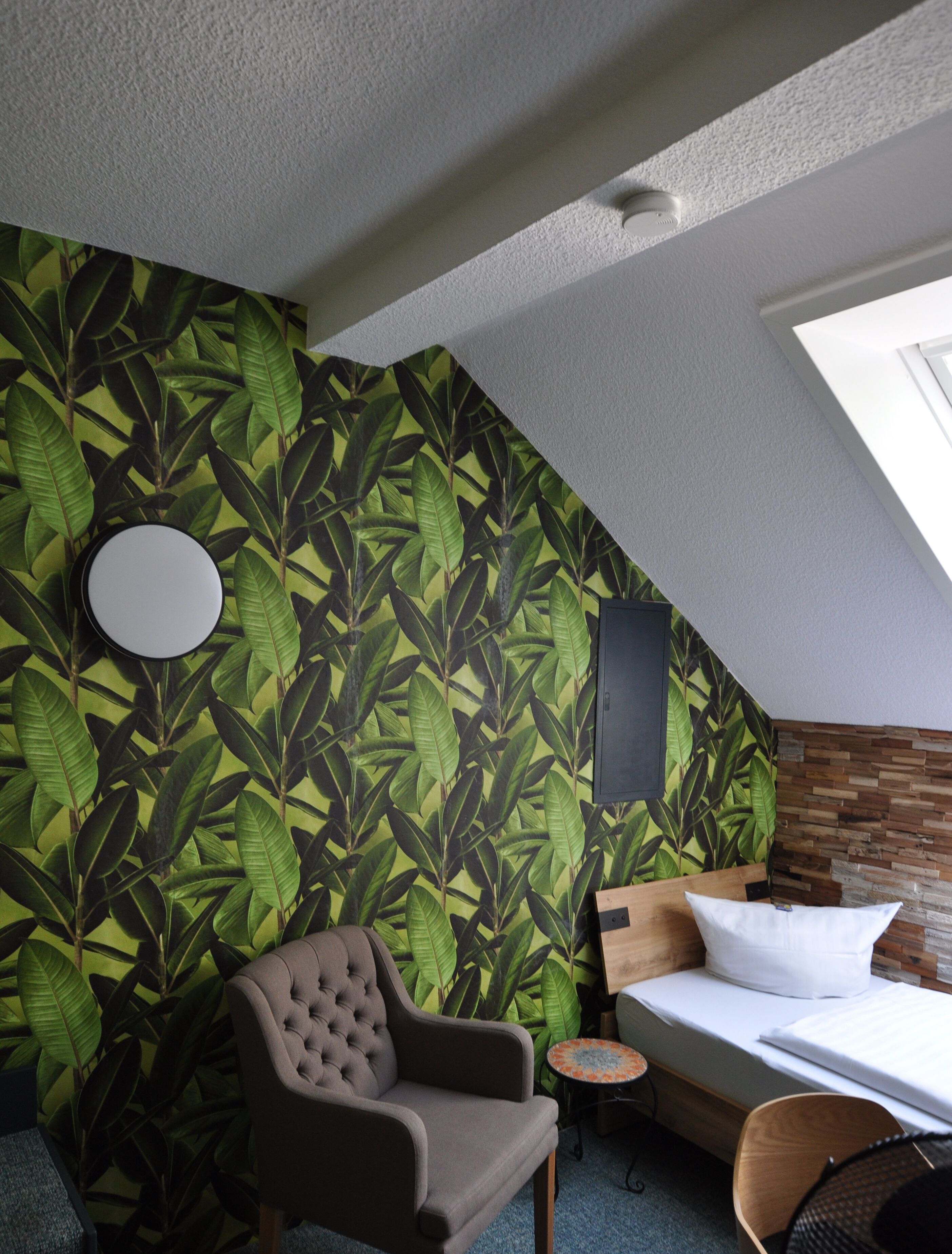 Een kamer met een schuine kap, een bed met een wit matras en kussen, een grijze fauteuil, een klein rond tafeltje en een muur met een bladpatroon.