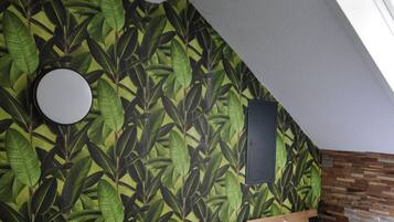 Een kamer met een schuine kap, een bed met een wit matras en kussen, een grijze fauteuil, een klein rond tafeltje en een muur met een bladpatroon.