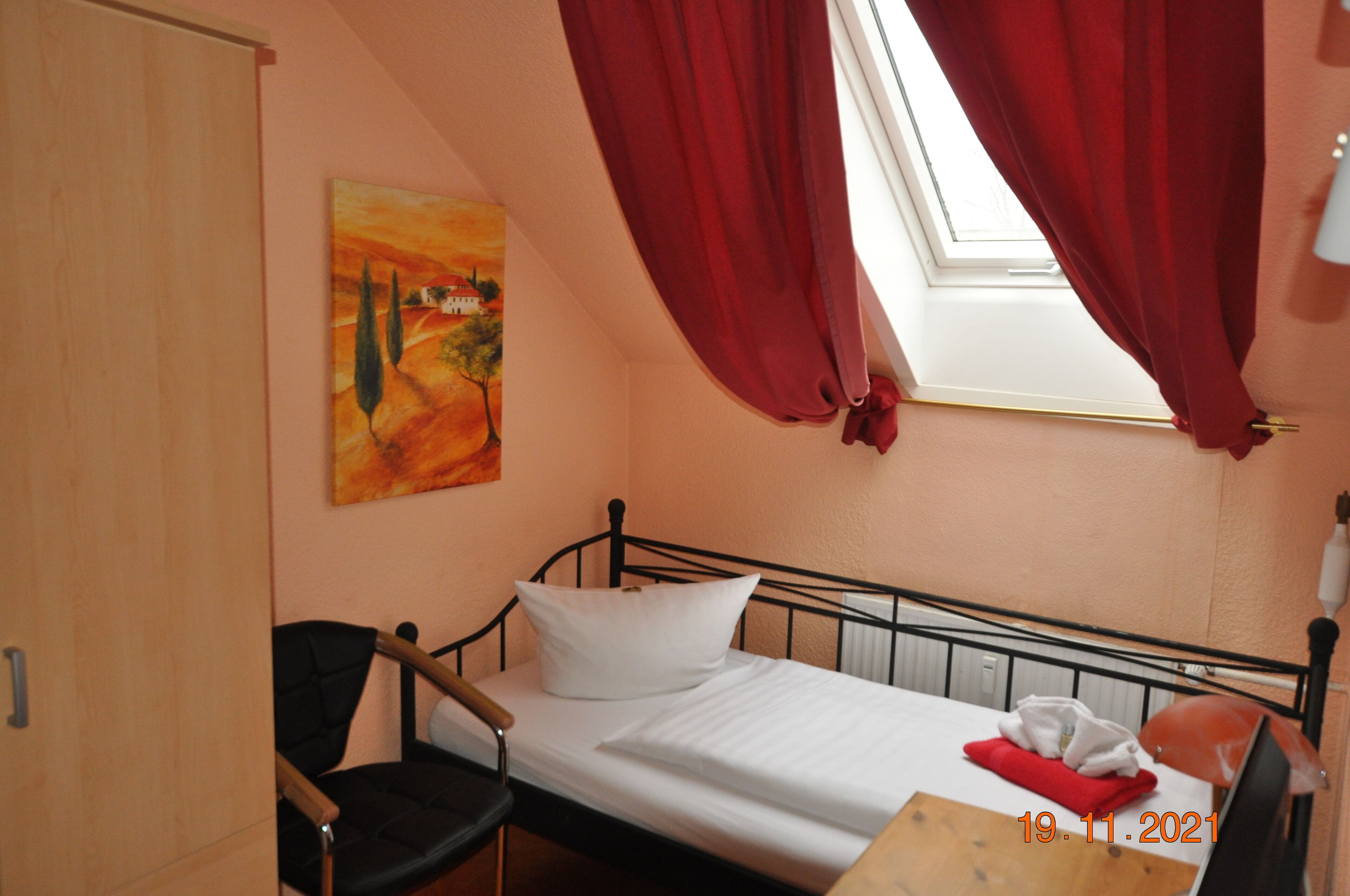Una cama individual con colchón y almohadas blancas, cabecera de metal negro, una mesa de madera, una silla negra, una ventana con cortinas rojas y un cuadro en la pared.