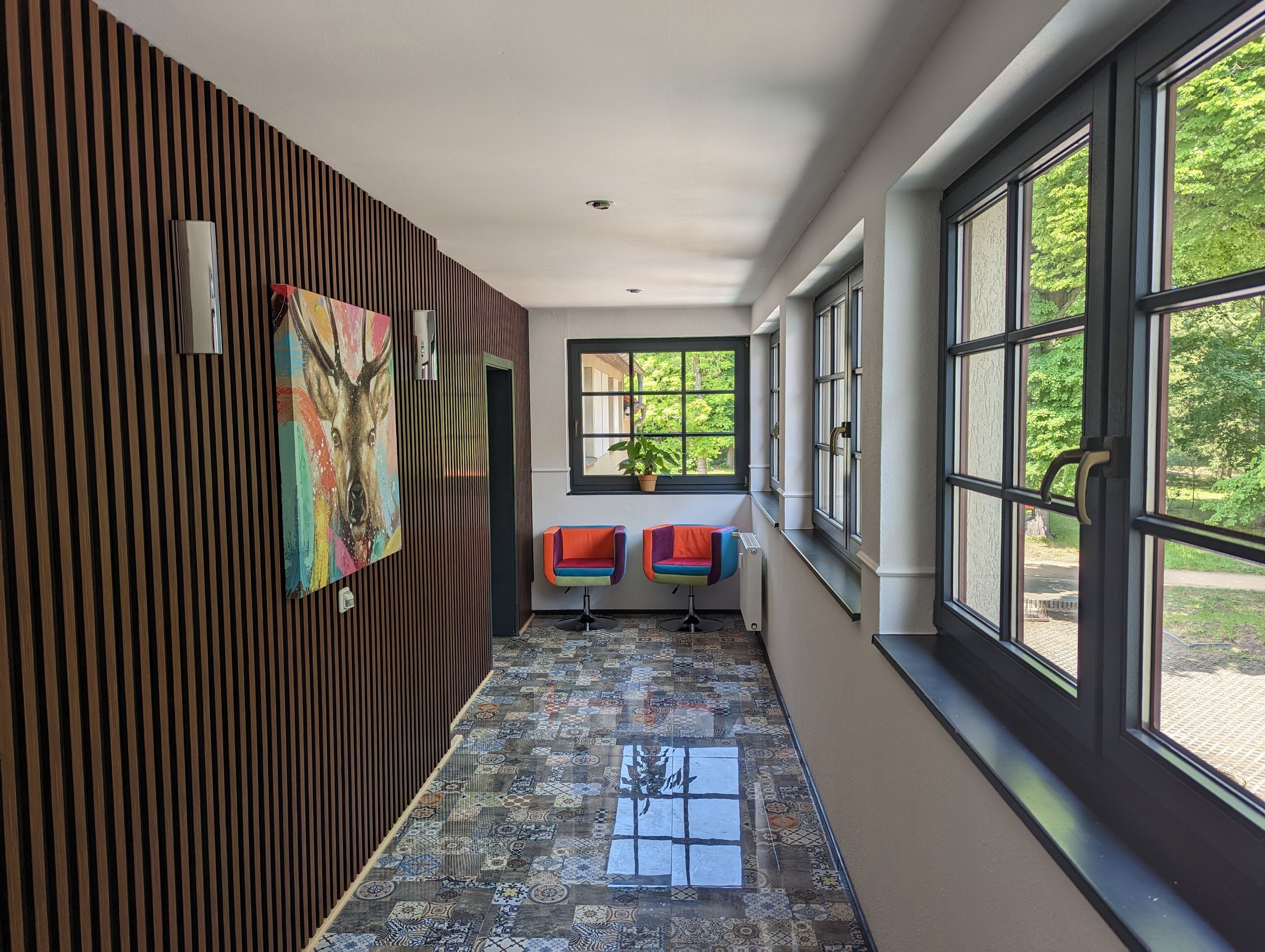 A hallway with a patterned floor, two colorful chairs, a large window with a view of greenery, and a wooden panel wall with a painting.