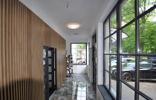 Un pasillo con pared de paneles de madera, suelo de baldosas y ventanas amplias que dan a la vista de coches aparcados y zonas verdes.