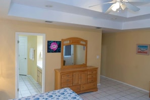 A bedroom with a wooden dresser, a ceiling fan, a bed with a patterned bedspread, and a door leading to another room. - Ground Floor Duplex * 1/2 block to Beach * BBQ ok (Blue Siren House) by RedAwning (South Padre Island)