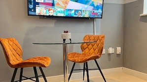 Une chambre moderne avec un téléviseur à écran plat fixé au mur, qui diffuse différents contenus. On y trouve deux fauteuils orange aux pieds noirs et une petite table entre les deux. La pièce est dotée d’un sol recouvert de moquette et d’un tapis à motifs.