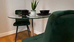 Un coin repas avec une table en verre, des chaises vert foncé, une plante en pot et une fenêtre avec des stores.
