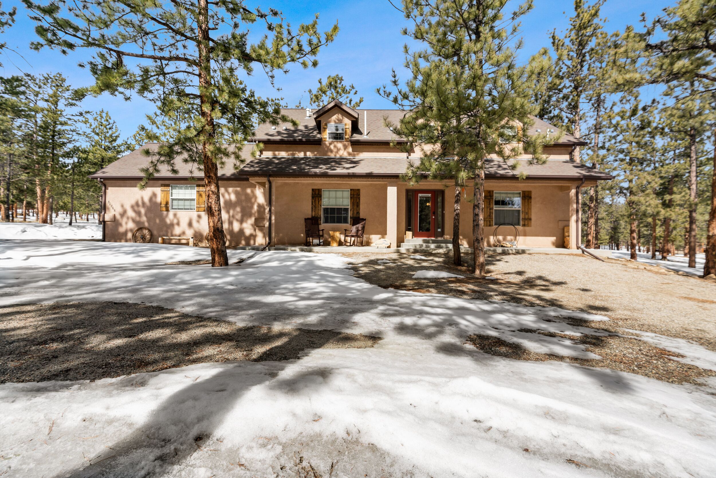 A two-story house with a front porch, surrounded by pine trees and covered in snow.
