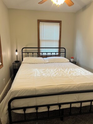 A bed with a headboard, a nightstand with a lamp, a window with blinds, and a ceiling fan.