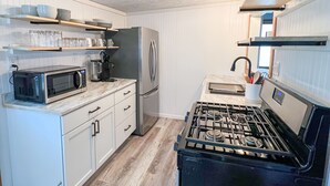 A compact kitchen with white cabinetry, a stainless steel refrigerator, a microwave, a gas stove, and wooden shelves above the counter. - Newly Remodeled Cabin Steps from Sylvan Lake (Rome City)