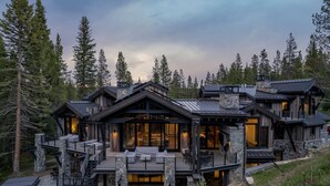 Une cabane en rondins à plusieurs étages, dotée de fondations en pierre, de grandes fenêtres et d’une terrasse avec des sièges d’extérieur.