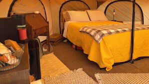 A tent with a bed, a small wooden table, a red cup, and a basket.