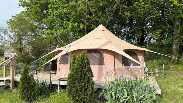 A beige tent with a platform and a staircase leading up to it, surrounded by greenery and trees.