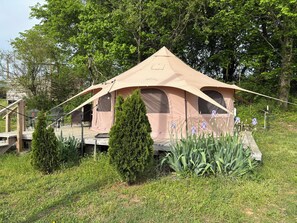 Een beige tent met een platform en een trap die erheen leidt, omringd door groen en bomen.