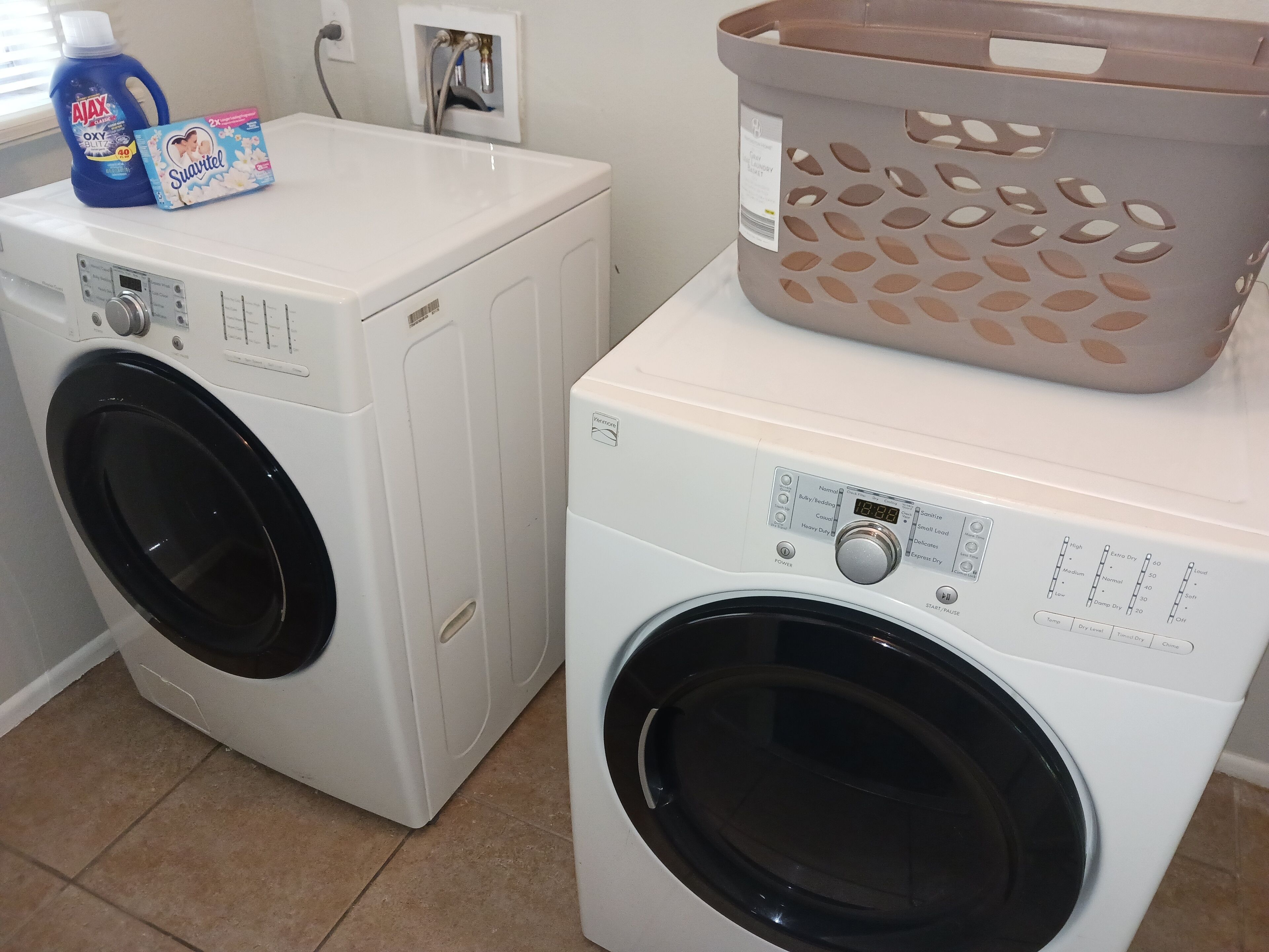 Two front-loading washing machines with control panels, a blue detergent bottle, and a gray laundry basket on top of one machine.