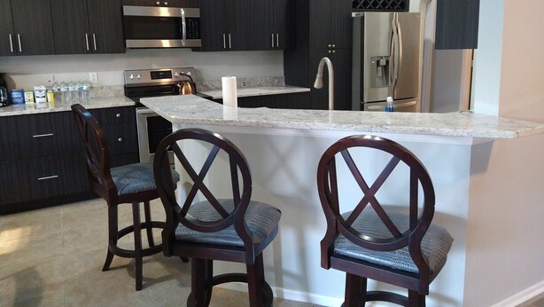 A modern kitchen with dark cabinets, stainless steel appliances, and a marble countertop. There are two wooden chairs with cushioned seats. - LOVE NEST--NEWLY RENOVATED Sleeps 7--CLOSE TO DOWNTOWN, AIRPORT & ATTRACTIONS (Orlando)