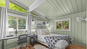 A bedroom with a bed, desk, chair, and a large window with curtains.