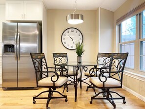 Une cuisine moderne avec un réfrigérateur en acier inoxydable, une table de salle à manger ronde entourée de quatre chaises, une horloge murale et un luminaire suspendu.
