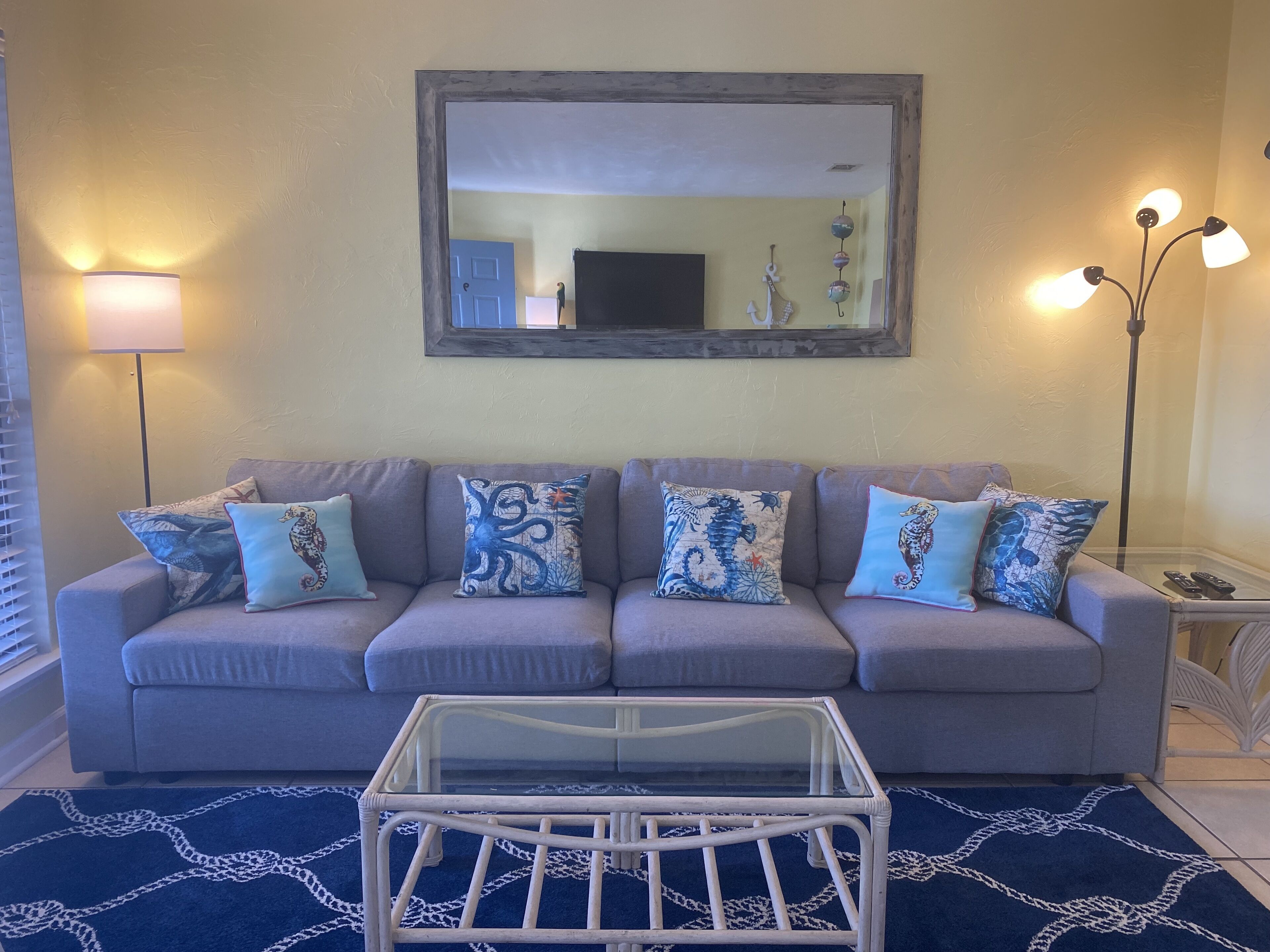 A living room with a grey sofa, patterned cushions, a glass coffee table, and a blue rug.