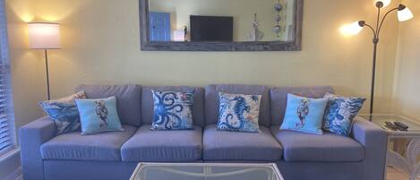A living room with a grey sofa, patterned cushions, a glass coffee table, and a blue rug.
