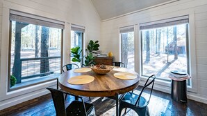 A dining area with a round wooden table, four chairs, a central woven basket, and large windows with blinds.