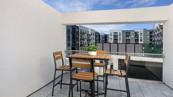 A balcony with a wooden table and chairs, a potted plant, and a view of modern apartment buildings.