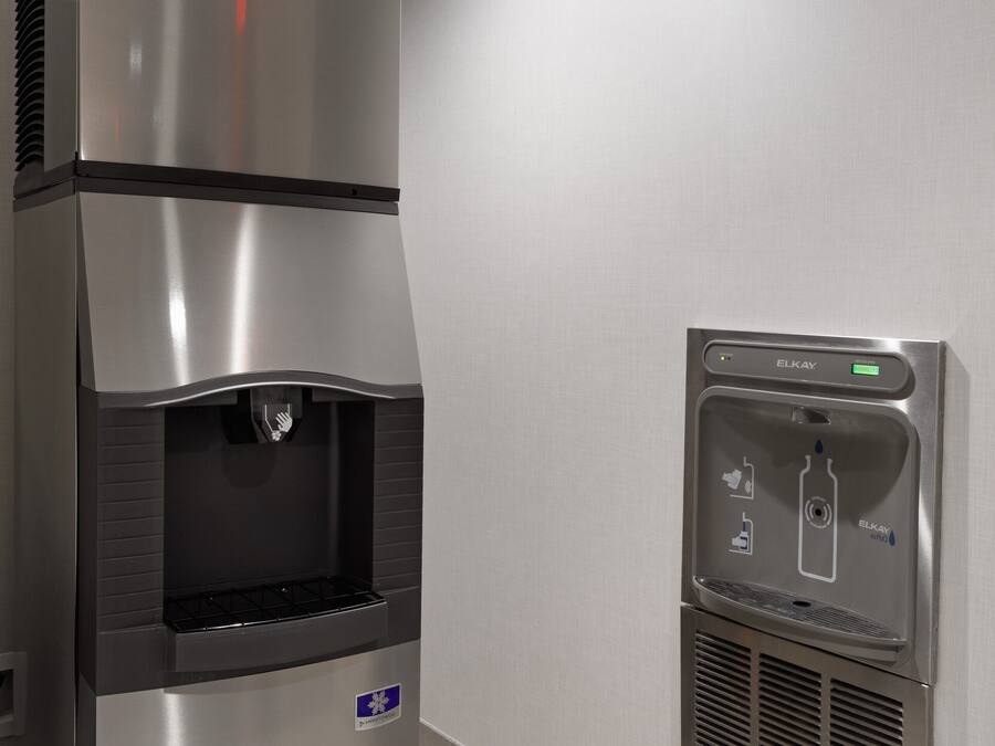 En vattenautomat i rostfritt stål med en digital display och en ikon som föreställer en vattenflaska, placerad bredvid en väggmonterad luftkonditioneringsenhet.