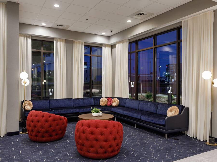 A modern hotel lobby with a dark blue herringbone floor, a dark blue sectional sofa, two red tufted ottomans, and a round coffee table with a plant.