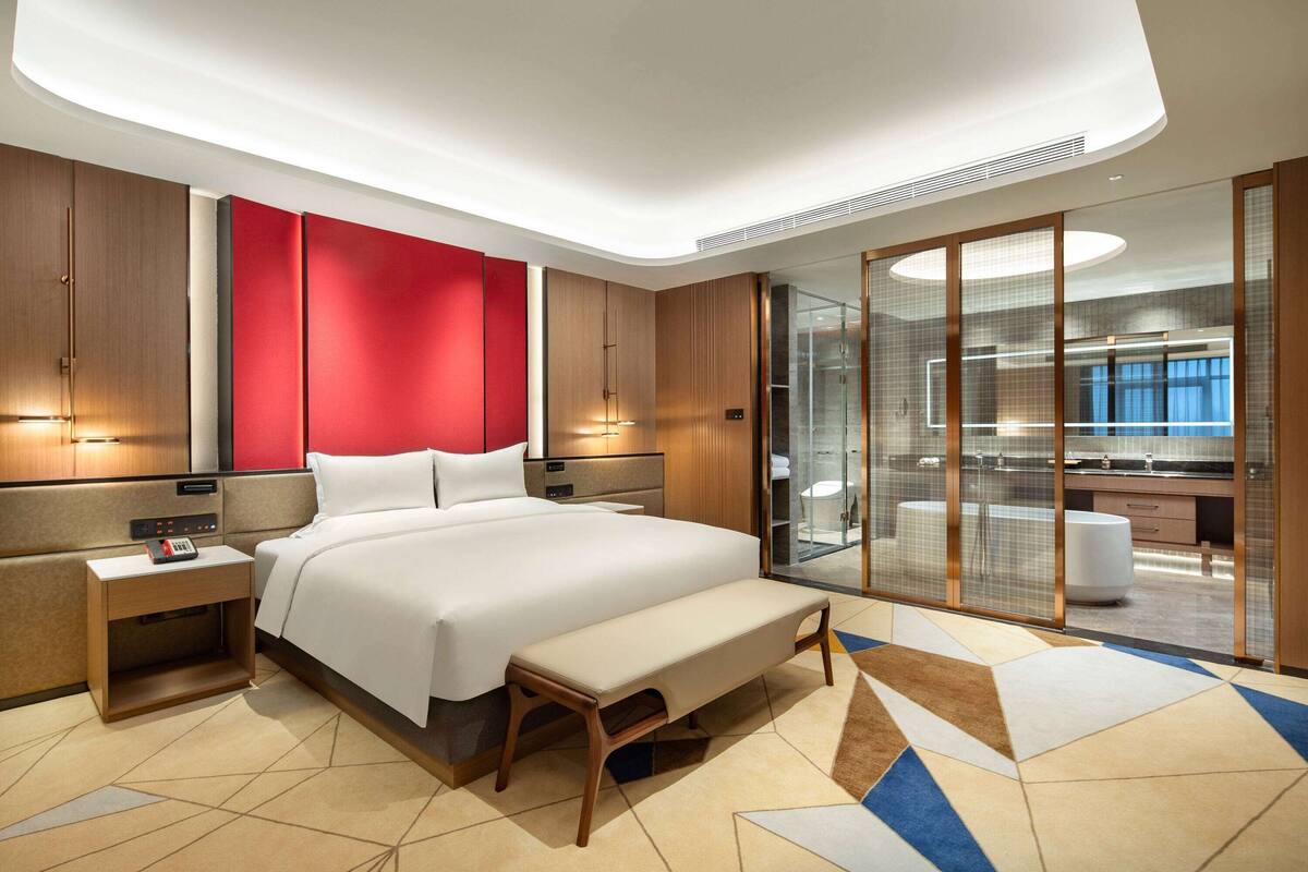 Ein modernes Hotelzimmer mit einem großen Bett, einem Nachttisch mit Telefon, einer Bank und einem durch eine Glasscheibe abgetrennten Badezimmer mit Badewanne.