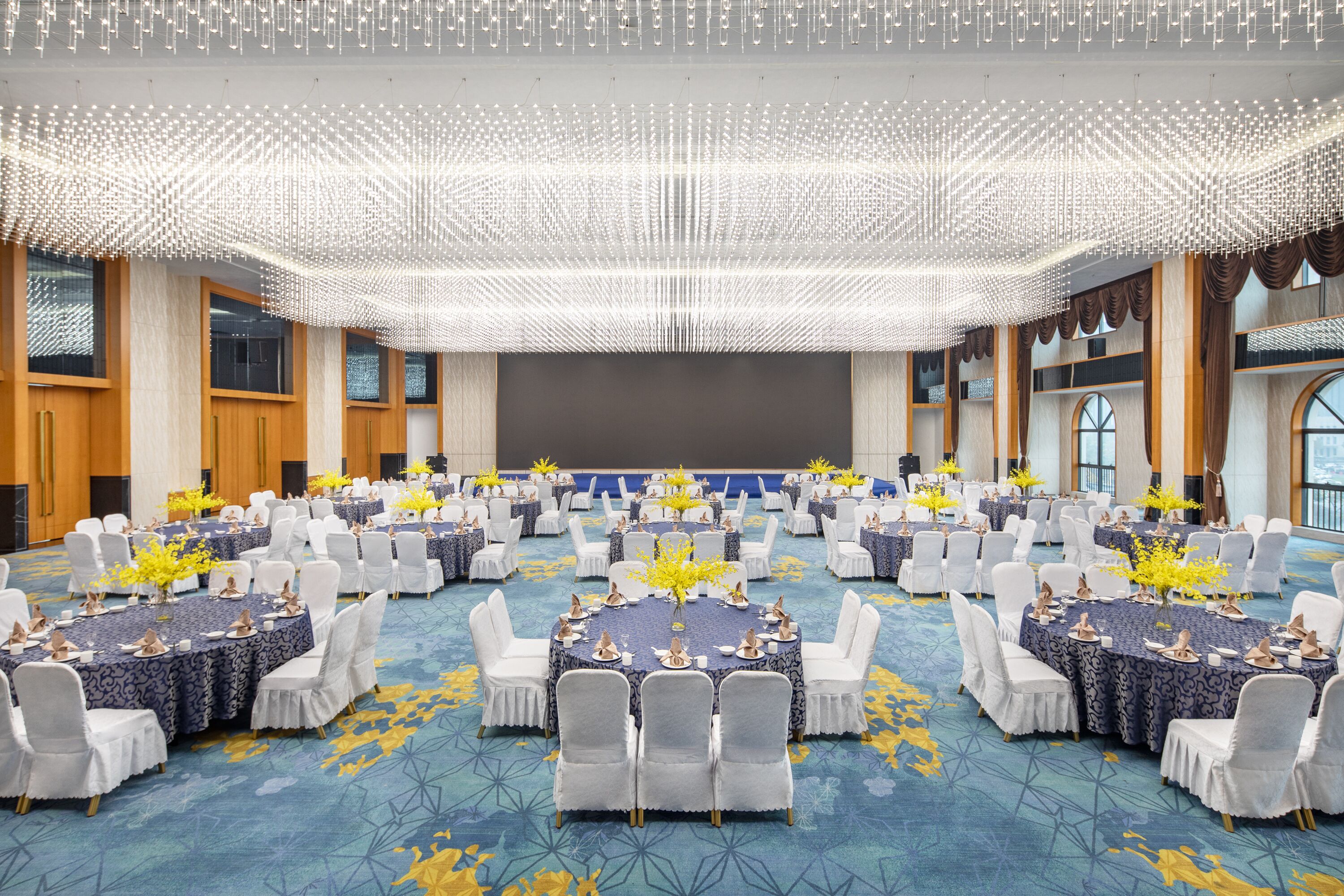 A large conference room with round tables and white chairs, decorated with yellow flowers and blue tablecloths.