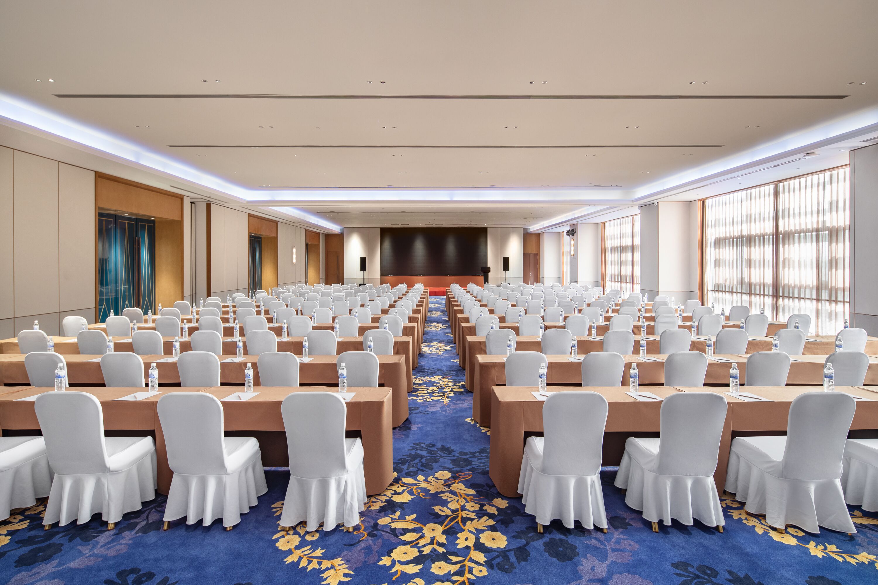 Un salón de conferencias con filas de mesas y sillas, un escenario al frente y ventanas grandes en un lateral.