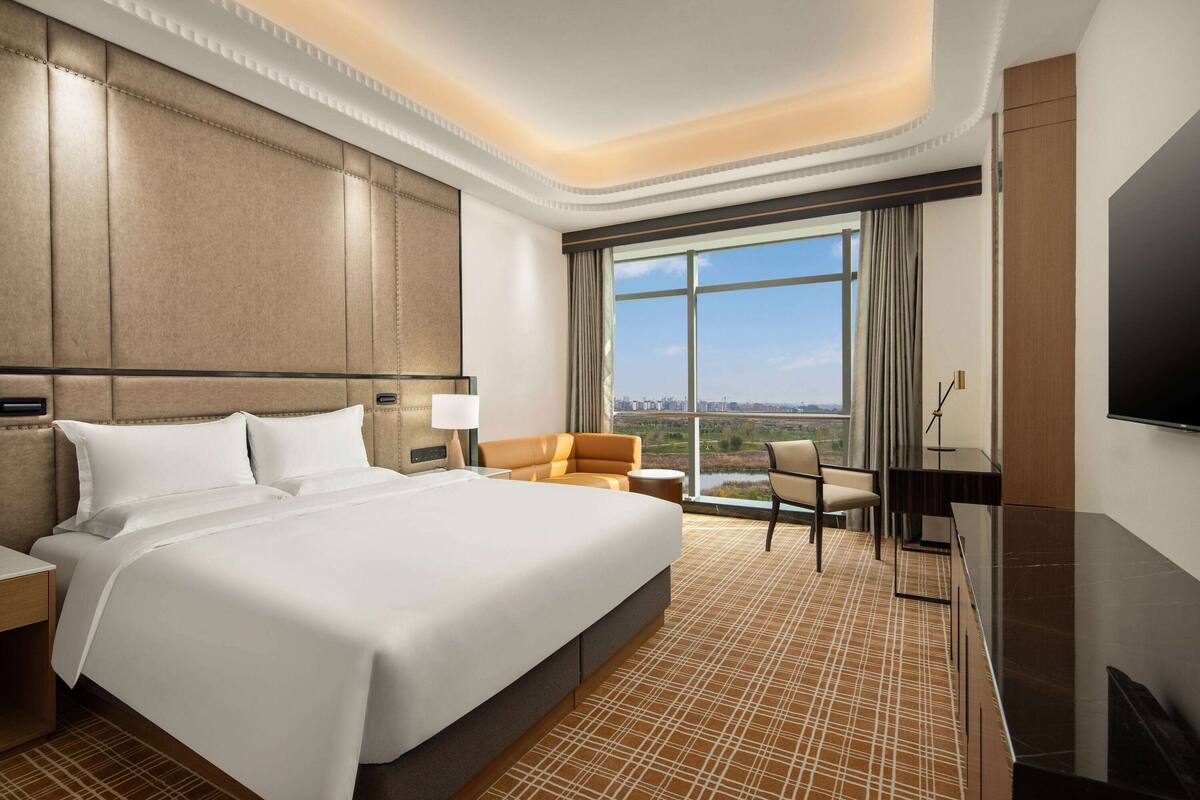 Ein Hotelzimmer mit einem großen Bett, einem Schreibtisch, einem Sessel, einem Fernseher und Blick auf die Stadt.