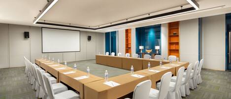 A conference room with a long table, white chairs, and a projector screen.