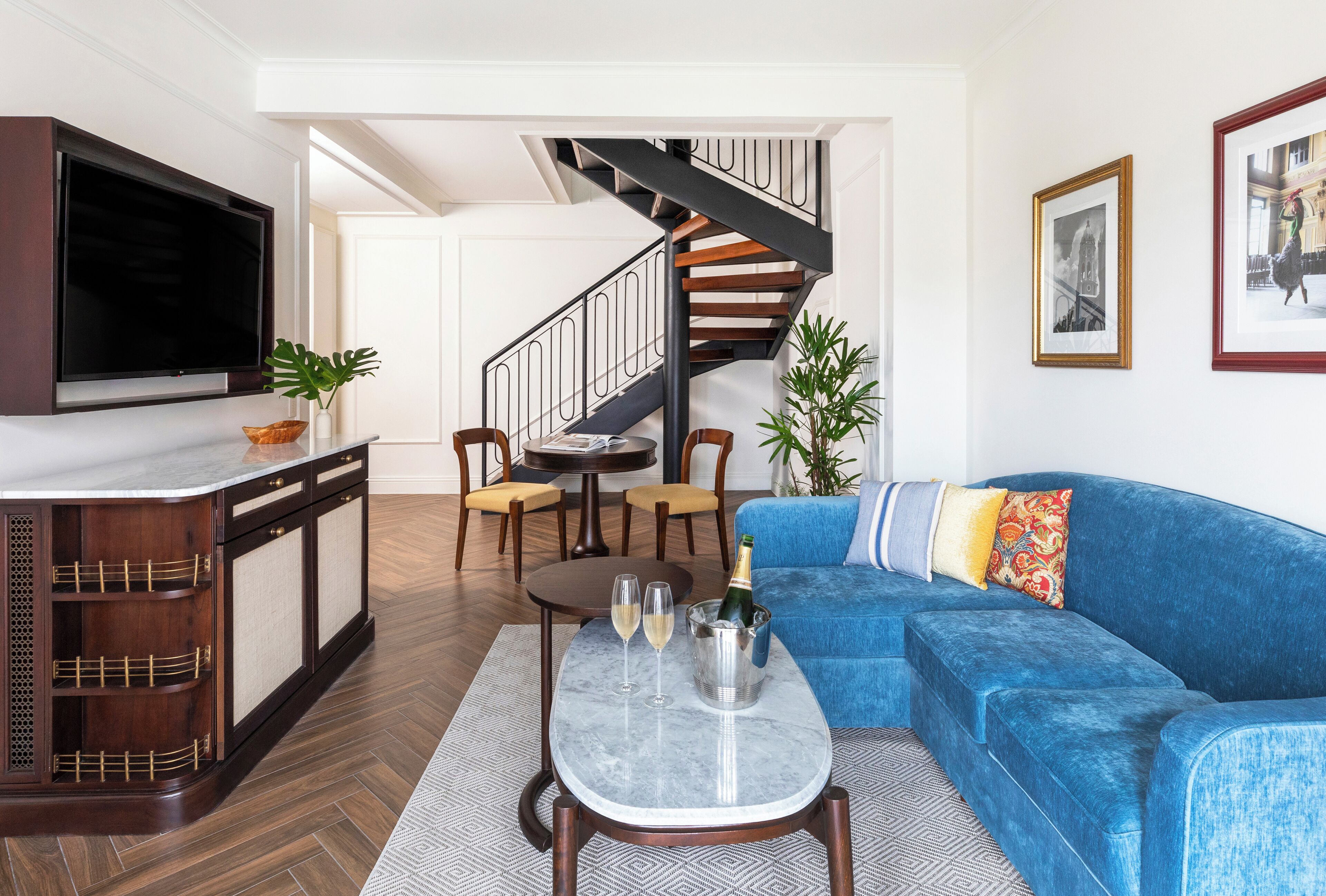 Una sala de estar con un sofá azul, un televisor montado en la pared, un mueble de madera, una mesa de comedor con sillas y una escalera.