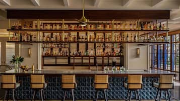 Zona de bar con varias estanterías llenas de botellas de licores, una barra central y cuatro taburetes de mimbre.